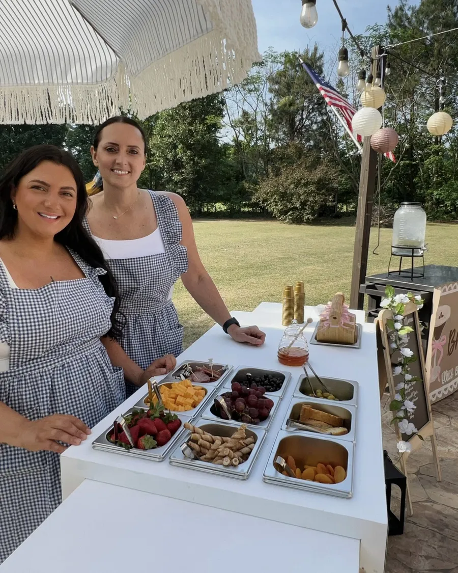 Karsen and Christina in matching blue gingham aprons, smiling behind a Bella Tavolo grazing cart